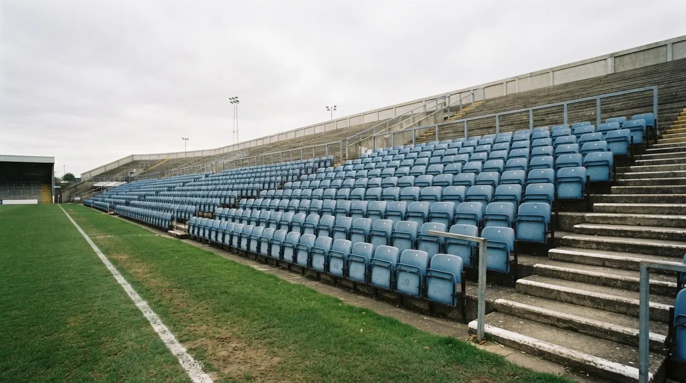 Rzędy pustych plastikowych krzesełek małego stadionu widziane z poziomu murawy pod pochmurnym niebem.
