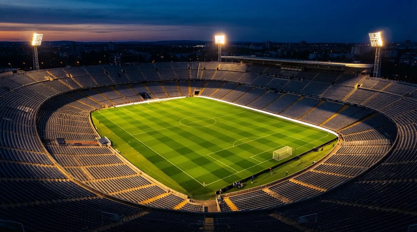 Widok z lotu ptaka na pusty stadion piłkarski o zmierzchu, murawa jaskrawozielona w świetle jupiterów.