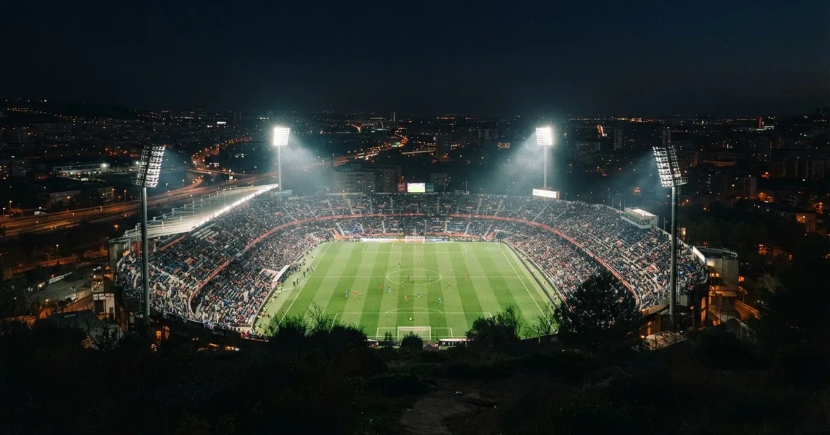 Stadion piłkarski nocą widziany z dużej odległości, jasne reflektory nad murawą i ciemne niebo wokół.