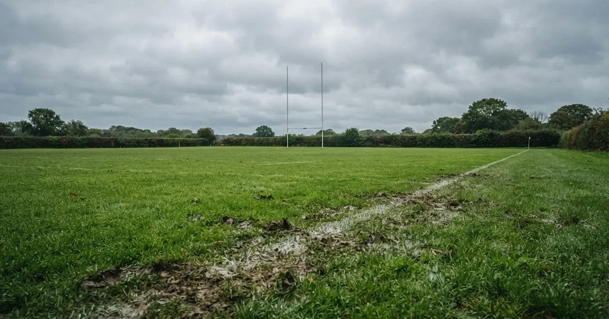 Boisko do rugby ze słupami bramki w oddali pod pochmurnym niebem, mokra trawa i ślady butów.
