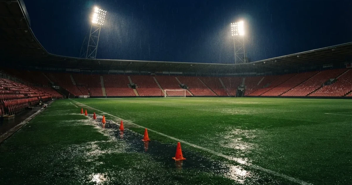 Pusty stadion piłkarski w deszczowy wieczór, mokra murawa odbija światła reflektorów, przy linii bocznej stoją pomarańczowe pachołki treningowe.