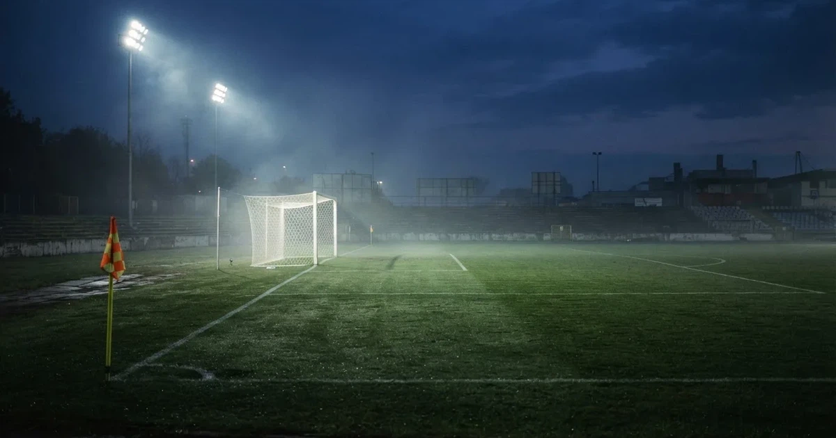 Pusta murawa stadionu miejskiego w chłodny jesienny wieczór, siatka bramki świeci w świetle reflektorów, nad boiskiem wisi mgła.