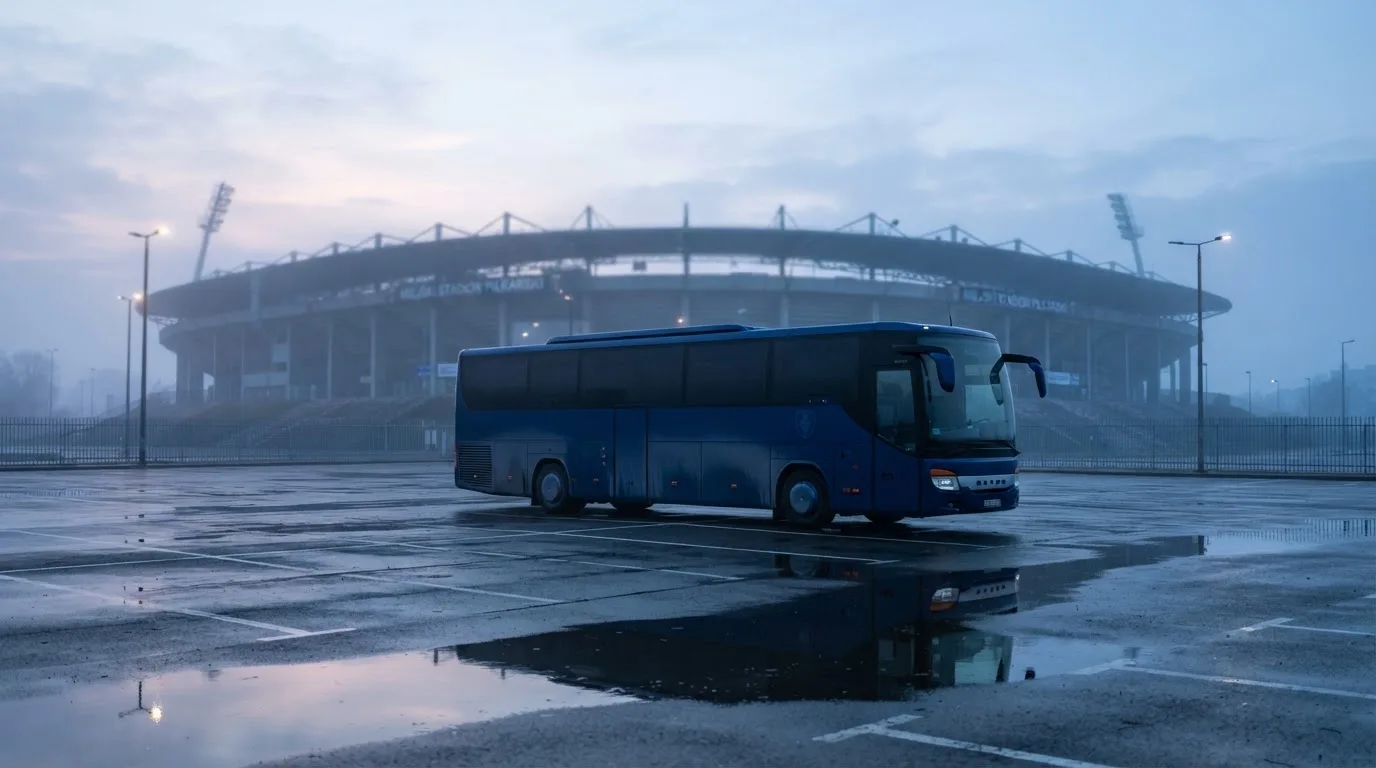 Autokar stoi na parkingu przed stadionem o świcie, rozmyte kontury trybun w tle, pusty plac i mokry asfalt.