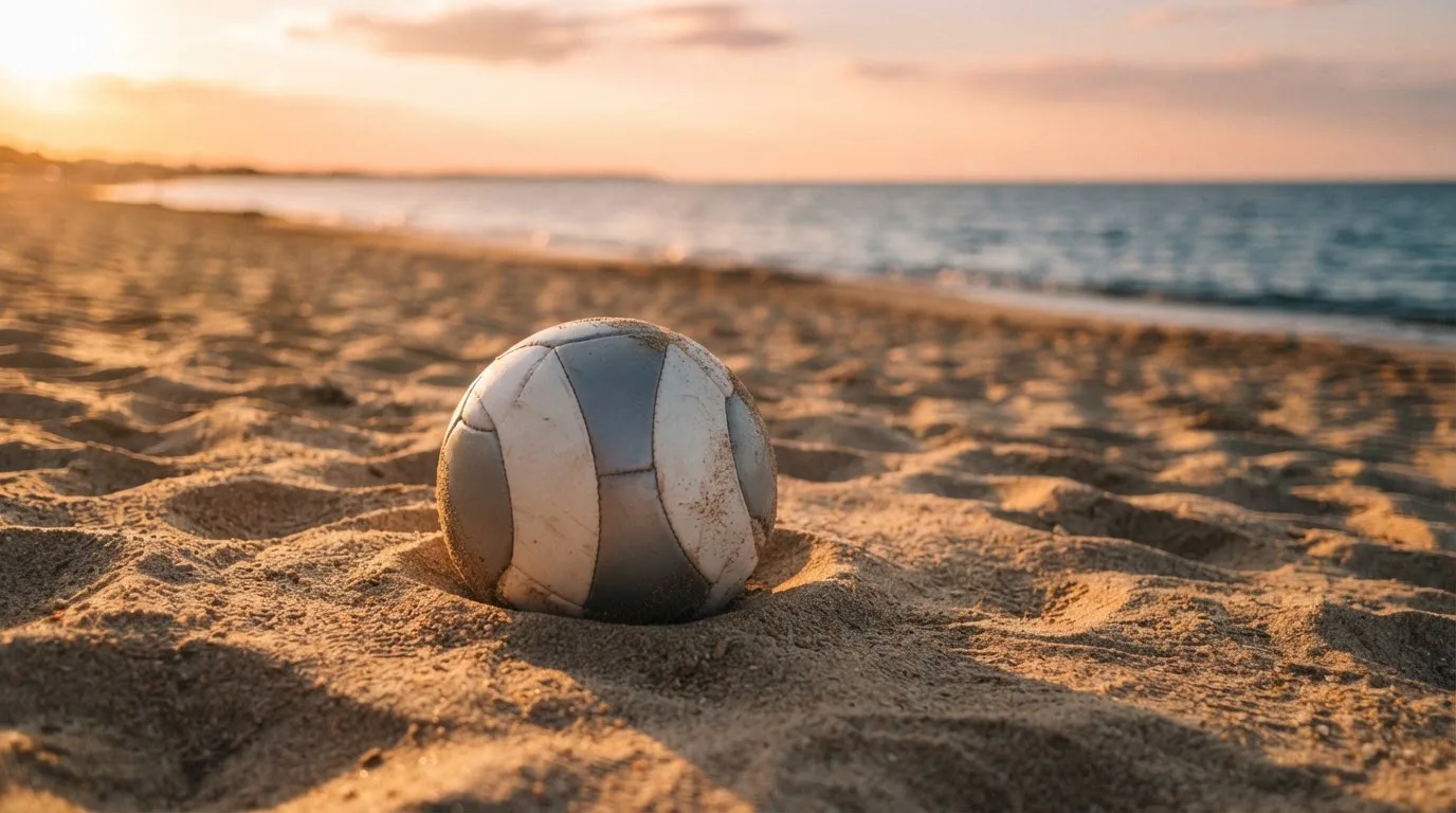 Piłka futbolowa leży na piasku boiska plażowego w ciepłym świetle zachodu, w tle morze i niski horyzont.