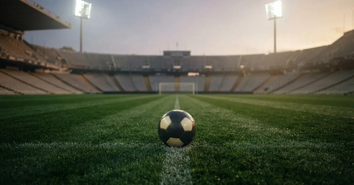 Piłka nożna leży na środku świeżo skoszonej murawy, w tle pusty stadion w świetle reflektorów wczesnym wieczorem.