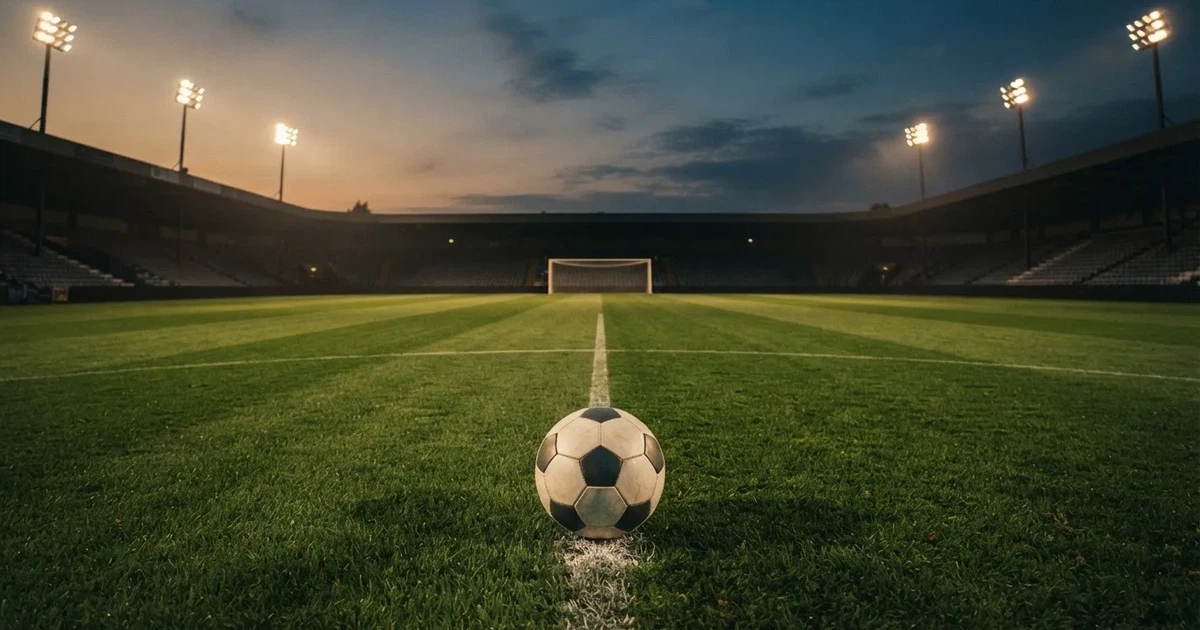 Piłka nożna leży na środku murawy dużego stadionu o zmierzchu, a nad boiskiem świecą rzędy reflektorów nad pustymi trybunami.